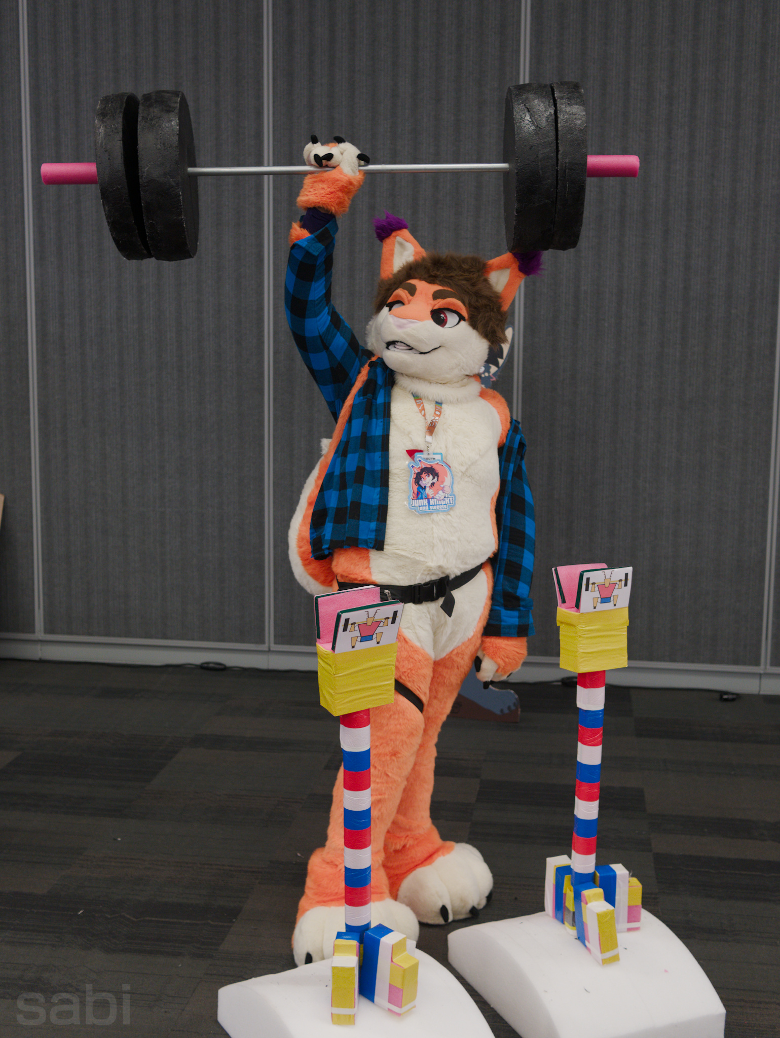a photo of a orange and white squirrel fursuiter holding a styrofoam dumbell above his head. his right leg is obscured by the styrofoam dumbell rack