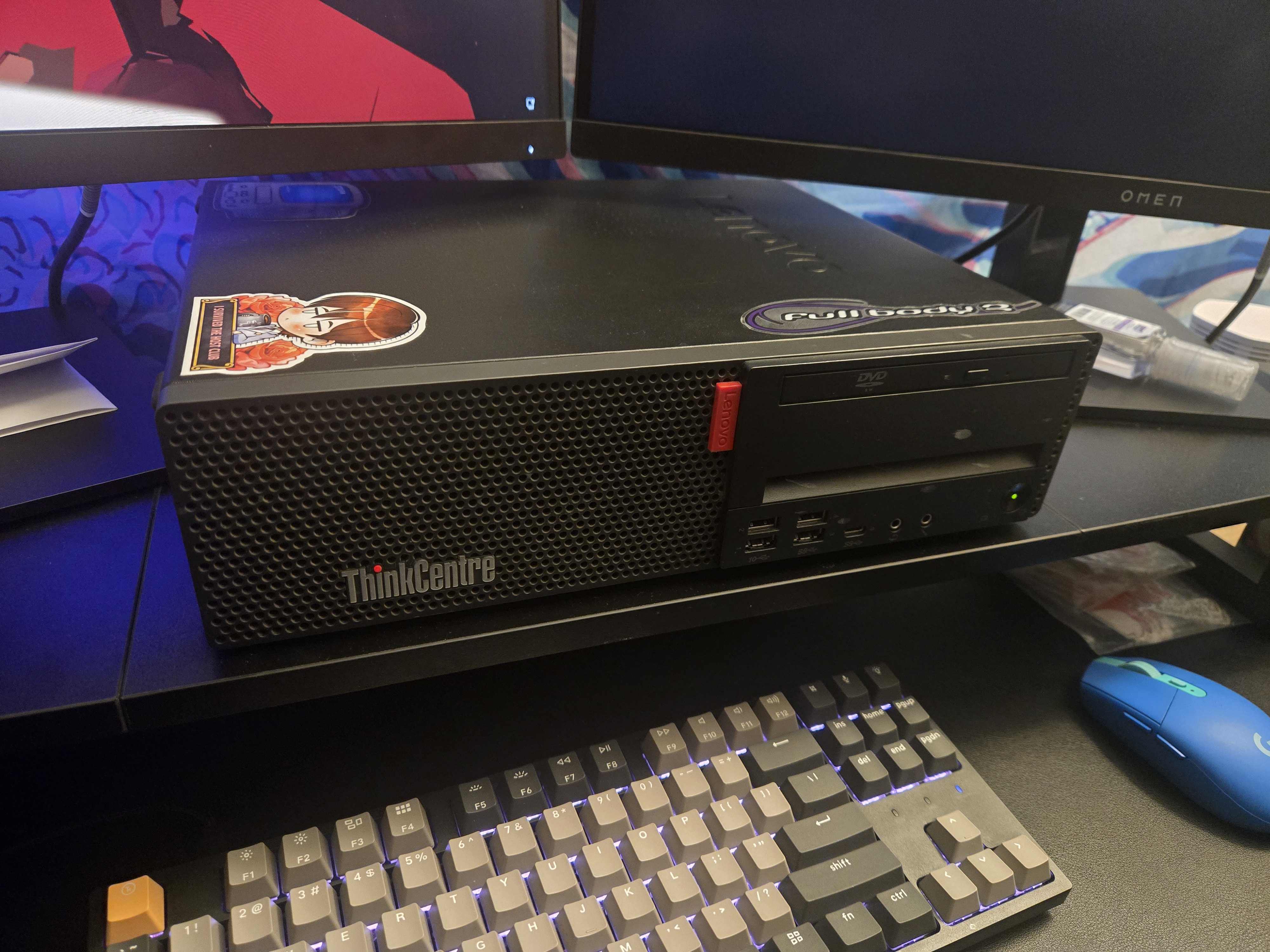 a powered-on black thinkcentre pc sits sideways on top of a black raiser on a desk sandwiched beneath and between two monitors