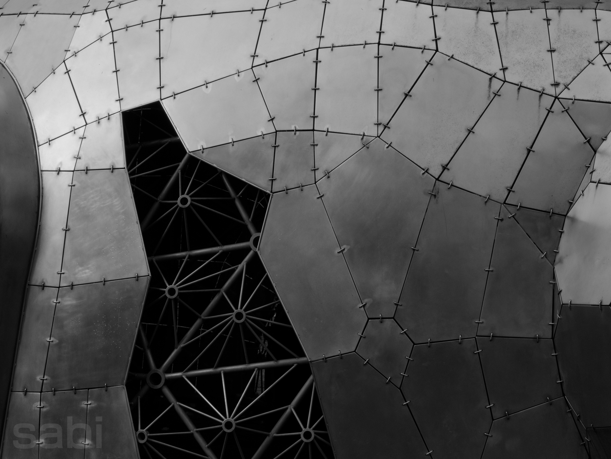 a monochrome photo of a silver sculpture with multiple conjoined panels, held up by bars, with what's almost a doorway leading inside